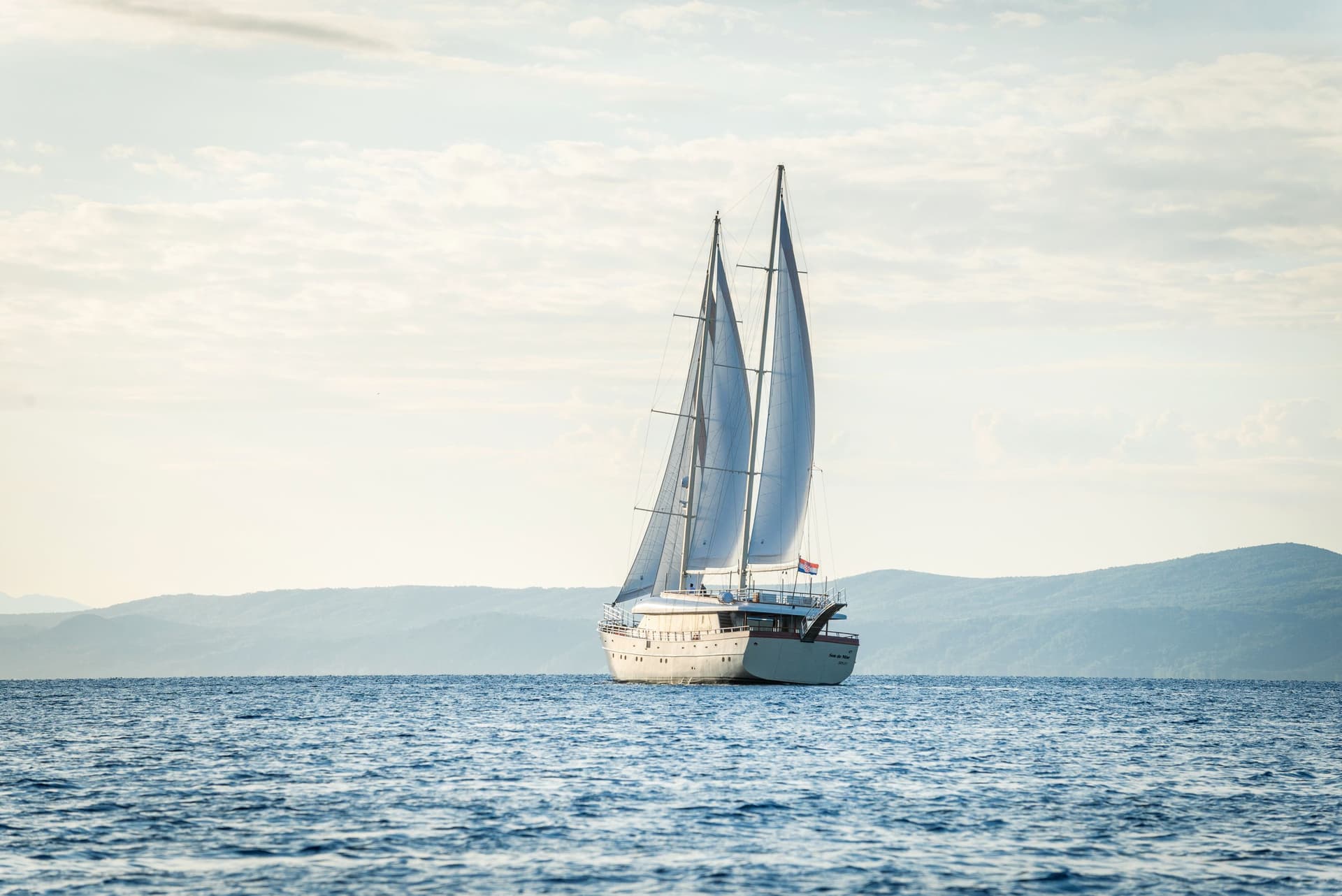 Large sailboat with sails up on blue sea near hazy mountains, Croatian flag visible.