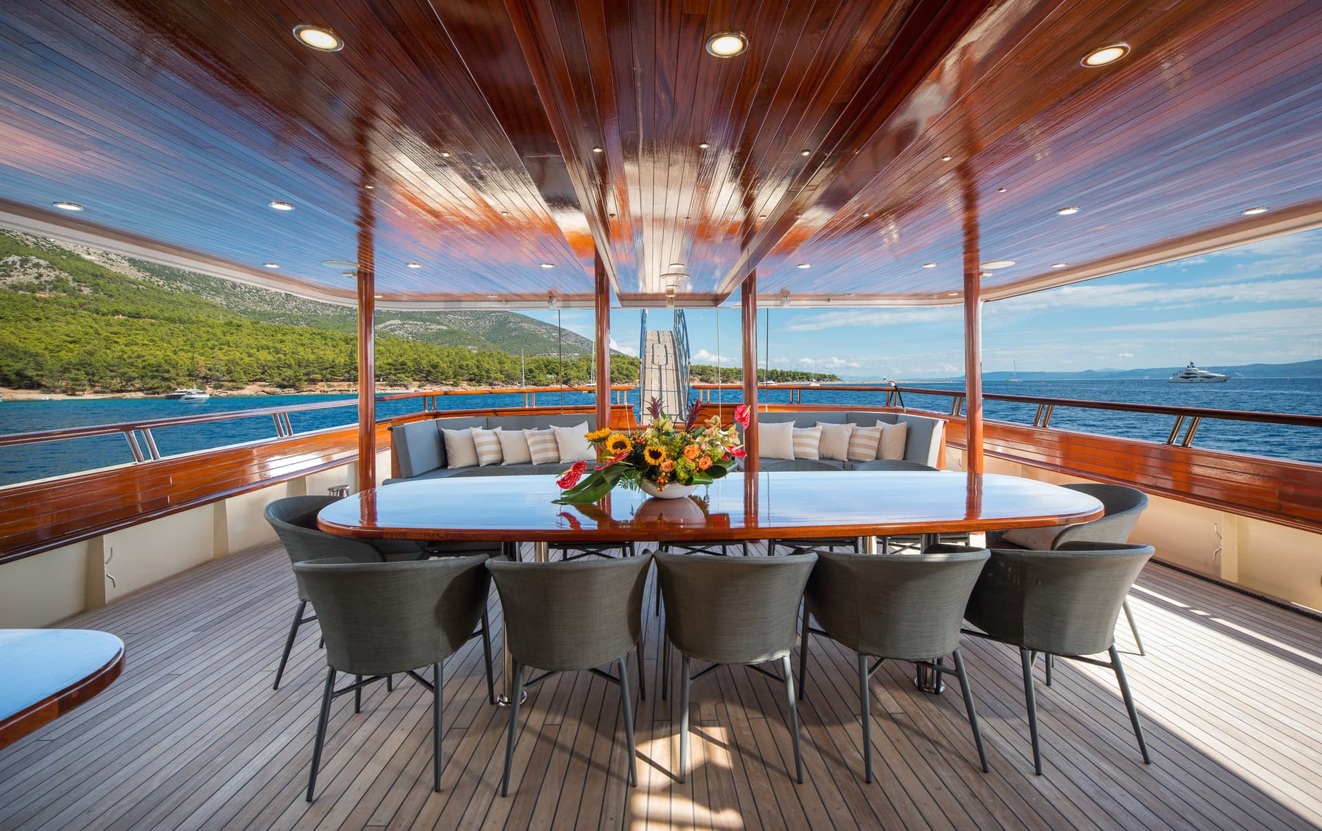 Outdoor dining area on a yacht with a wooden table, chairs, and view of a forested coastline.