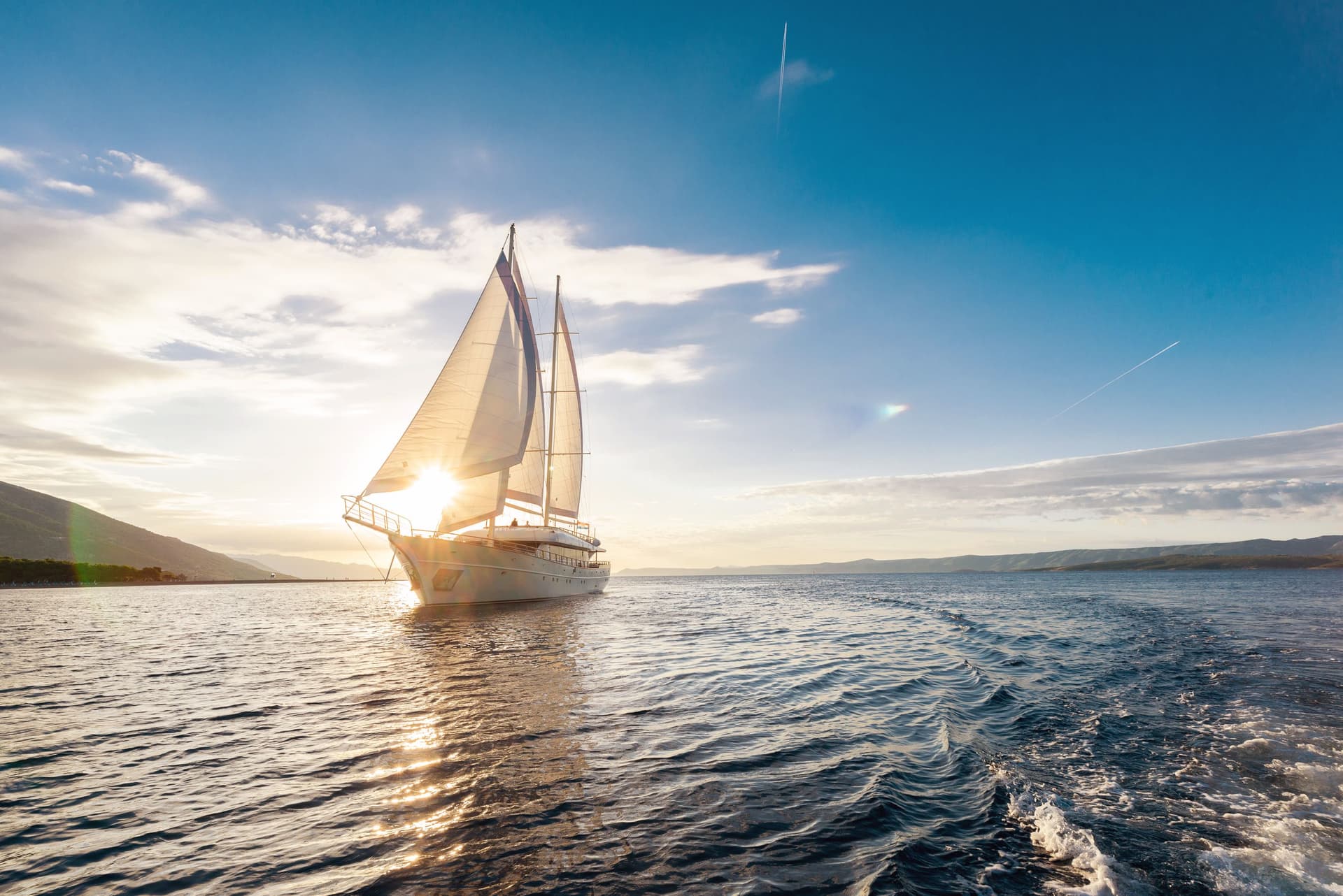 Large sailboat with sails up on calm sea water near mountains at sunset