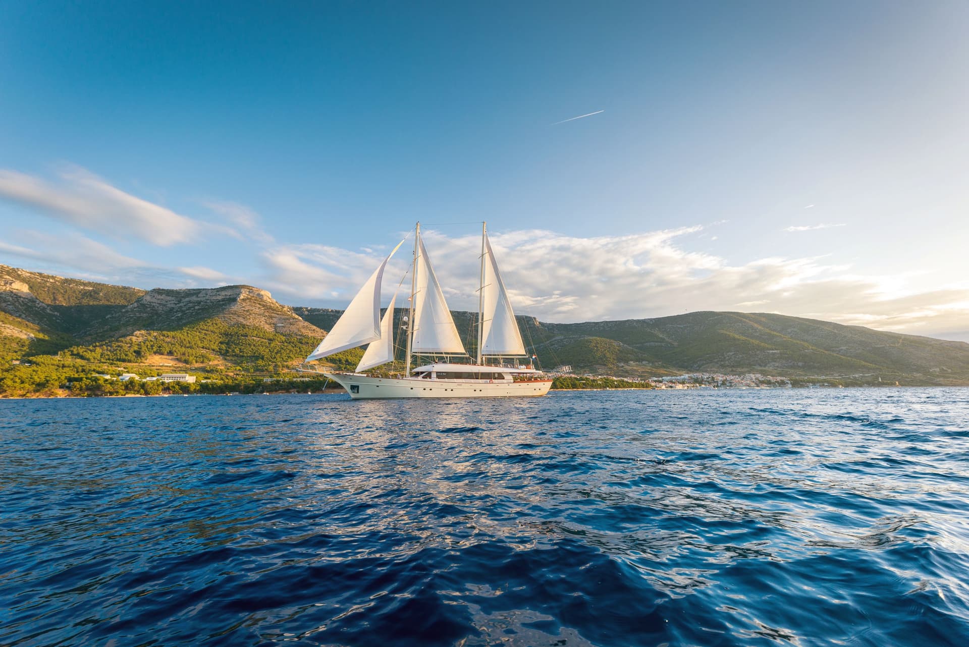 Large sailboat with white sails on deep blue water near a mountainous coastline.