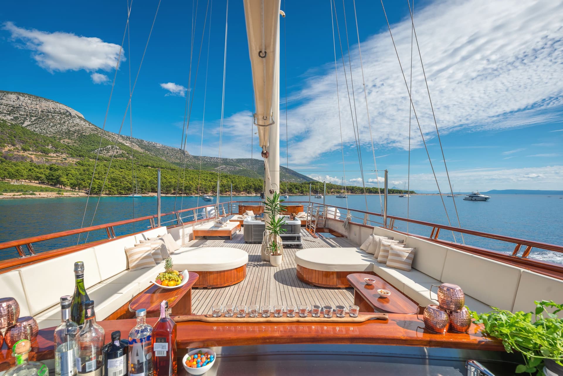 Spacious yacht deck with bar, lounge, and hot tub near forested coast under blue sky.