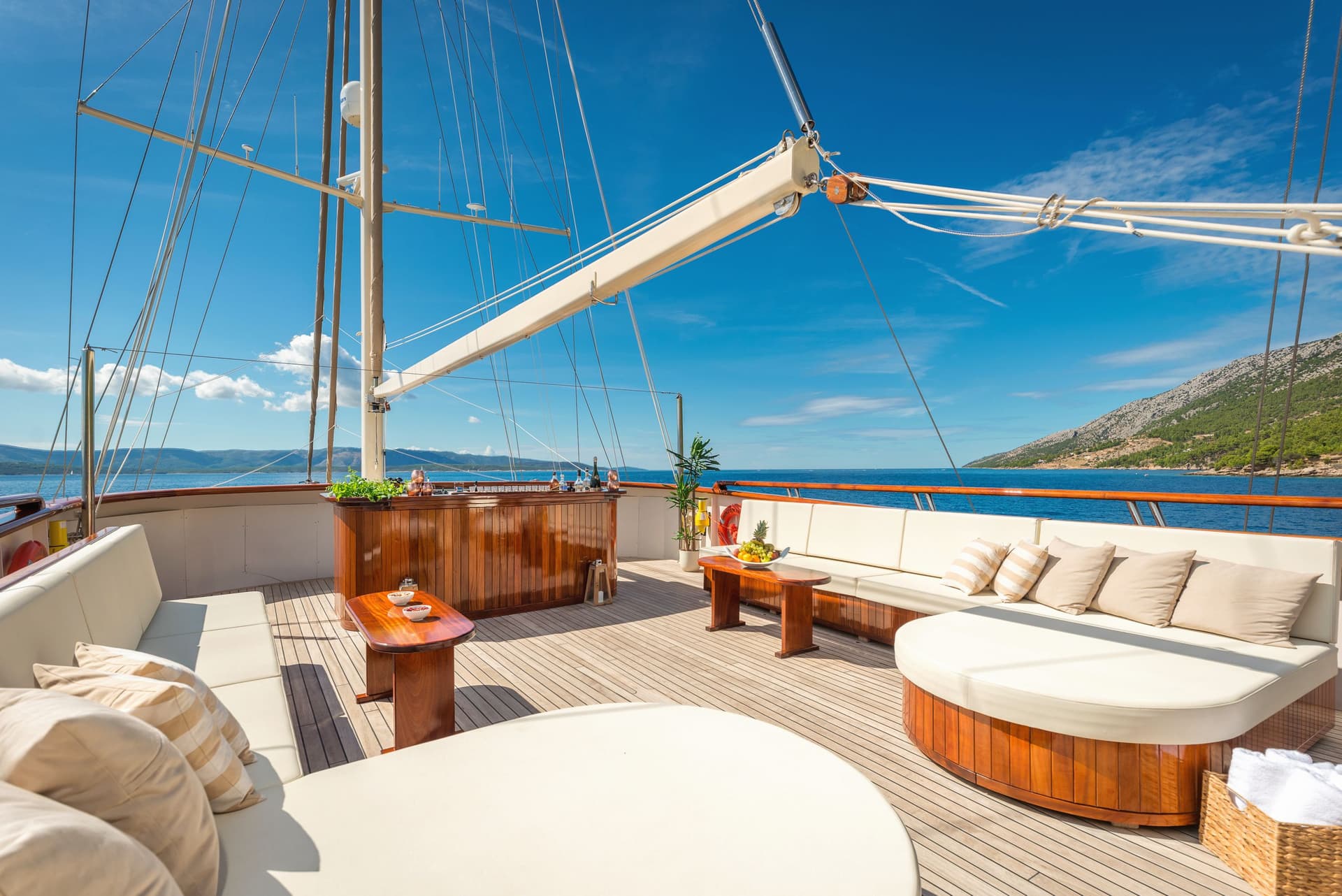 Sundeck lounge area with wooden bar and seating on a yacht near a green, rocky coastline.