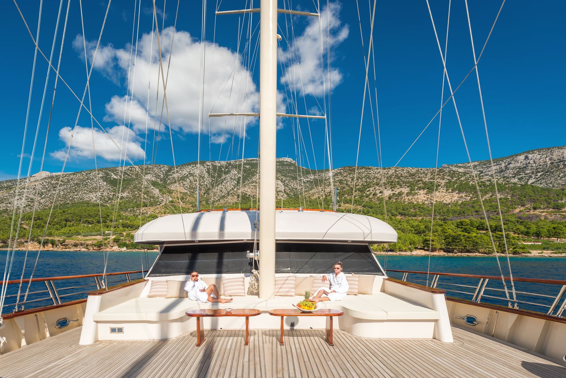 Two people relaxing on the deck of a yacht near a forested, rocky coastline.