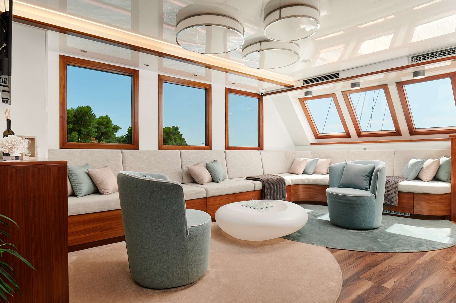 Modern yacht lounge with built-in seating, light blue armchairs, and views of green trees.