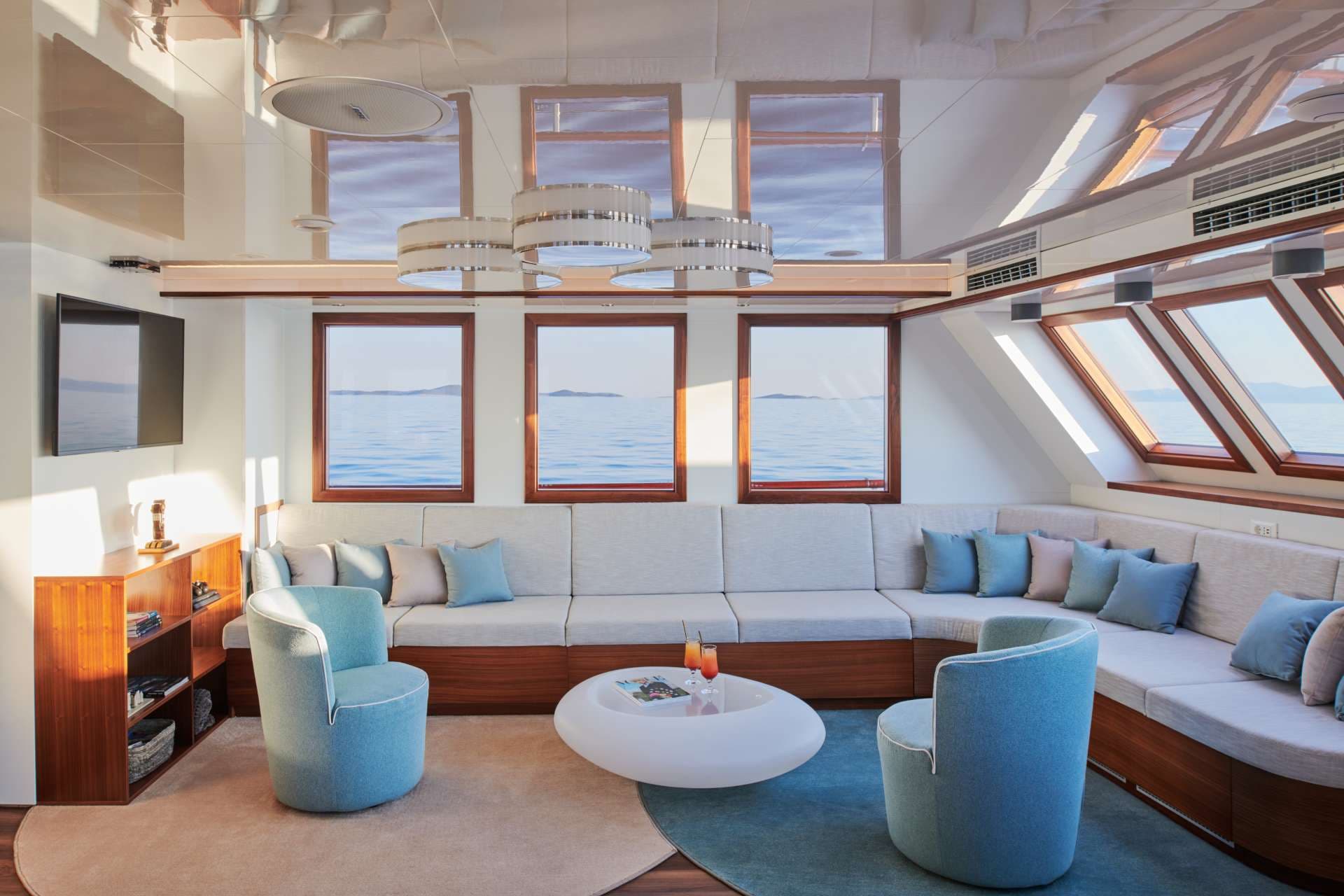 Lounge area on a boat with sea view windows, light blue chairs, and cocktails on a white table.
