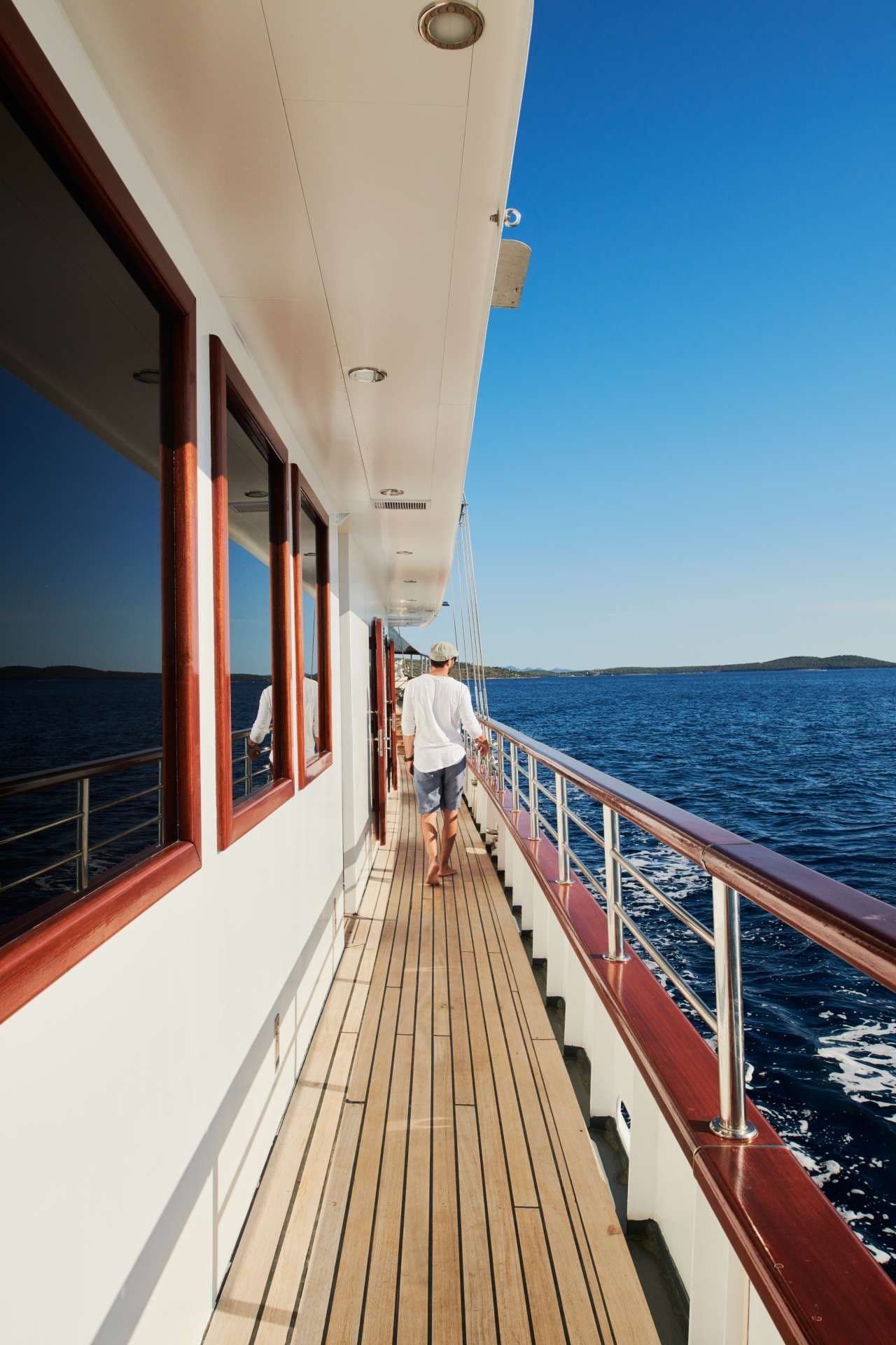 Man walking on teak deck of boat past windows on sunny day near islands.
