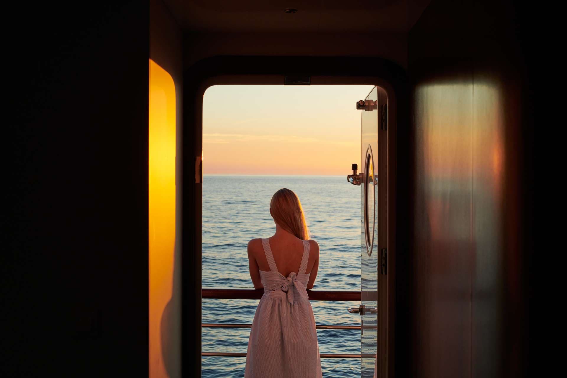 Woman in dress looking at sunset over the sea from a boat doorway