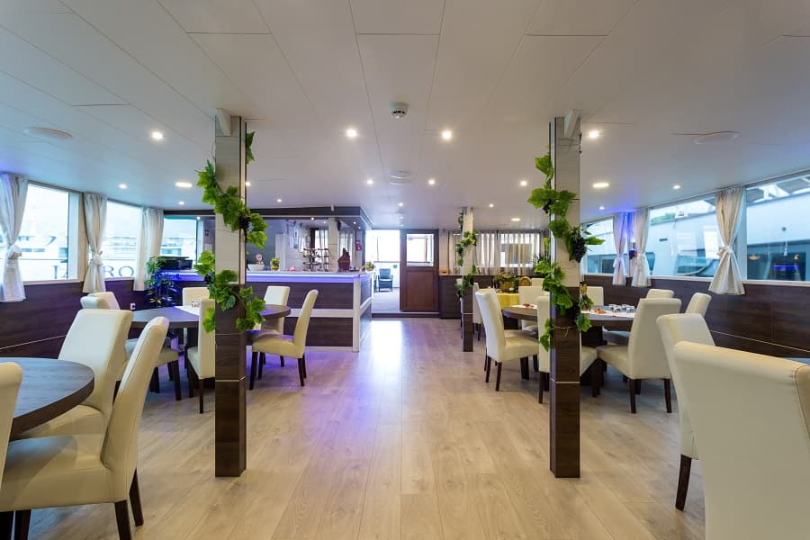 Dining room interior with white chairs, wooden tables, and grape vine decor on pillars, likely on a boat.