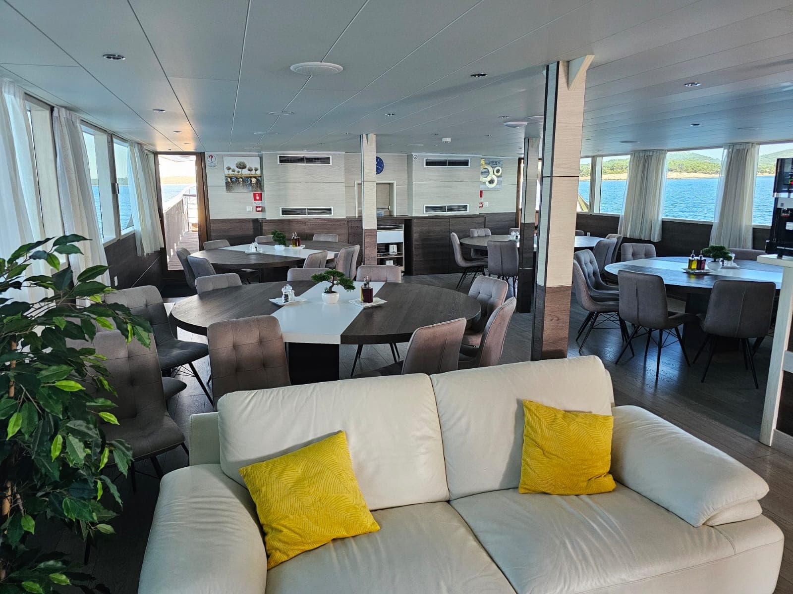 Lounge area with dining tables on a boat, overlooking blue water and green hills.
