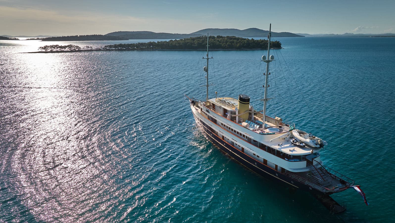 Large yacht anchored in blue coastal waters near forested islands under bright sun.