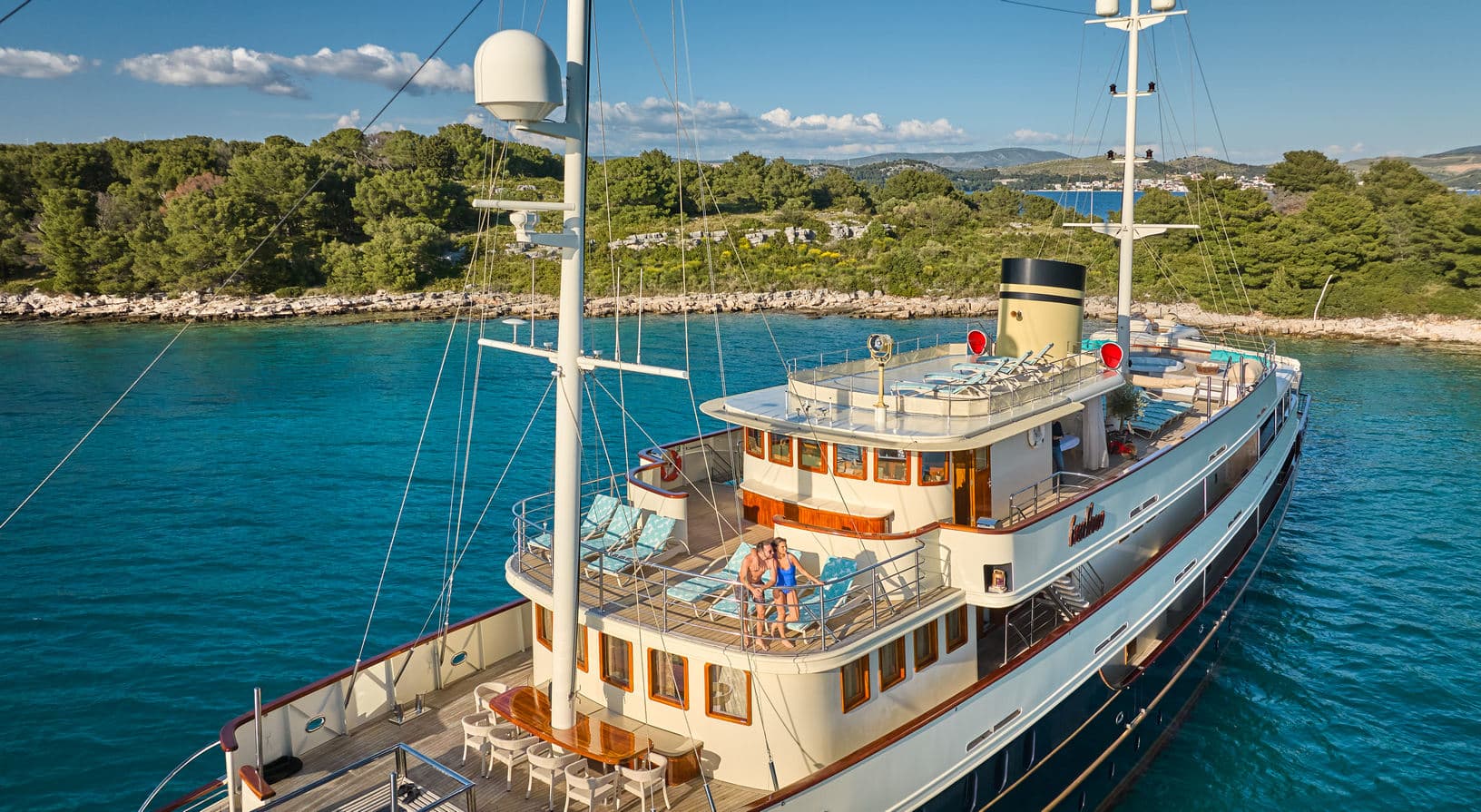 Luxury yacht anchored in turquoise coastal waters near a wooded island shore.