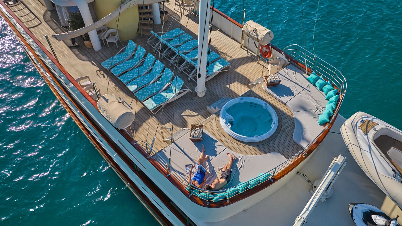 Yacht deck with hot tub, sun loungers, and two people sunbathing over turquoise water.