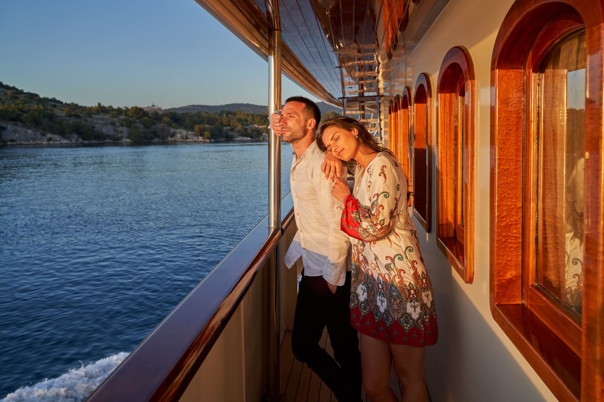 Couple enjoying sunset view from a yacht deck overlooking a hilly coastline.