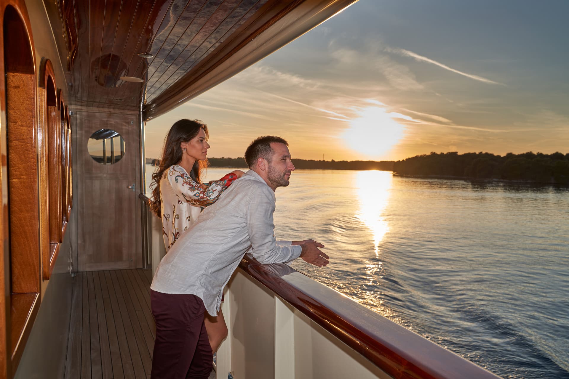 Couple watching sunset over water from the deck of a yacht with polished wood trim.