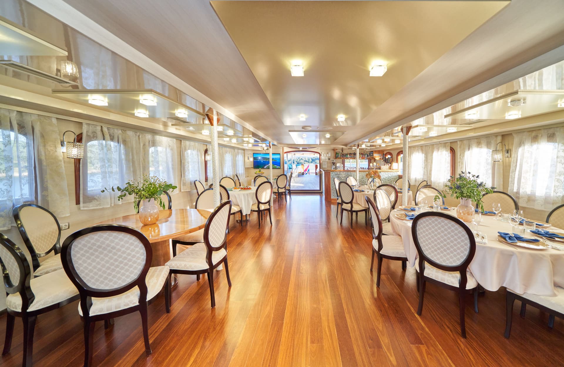 Dining salon interior on a boat with wood floors, set tables, and an open stern view.