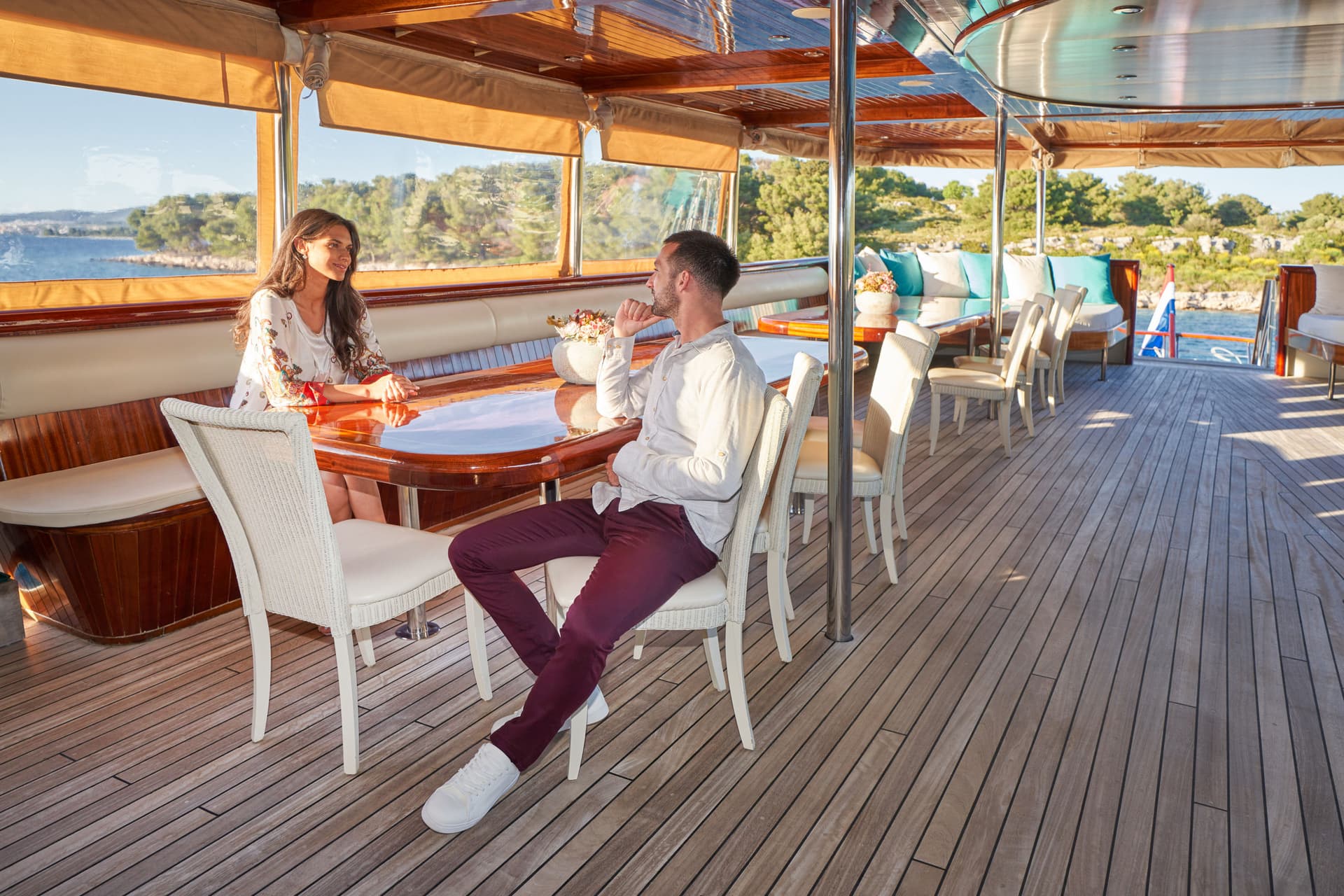 Couple dining on teak deck of a boat with coastal scenery visible through windows.