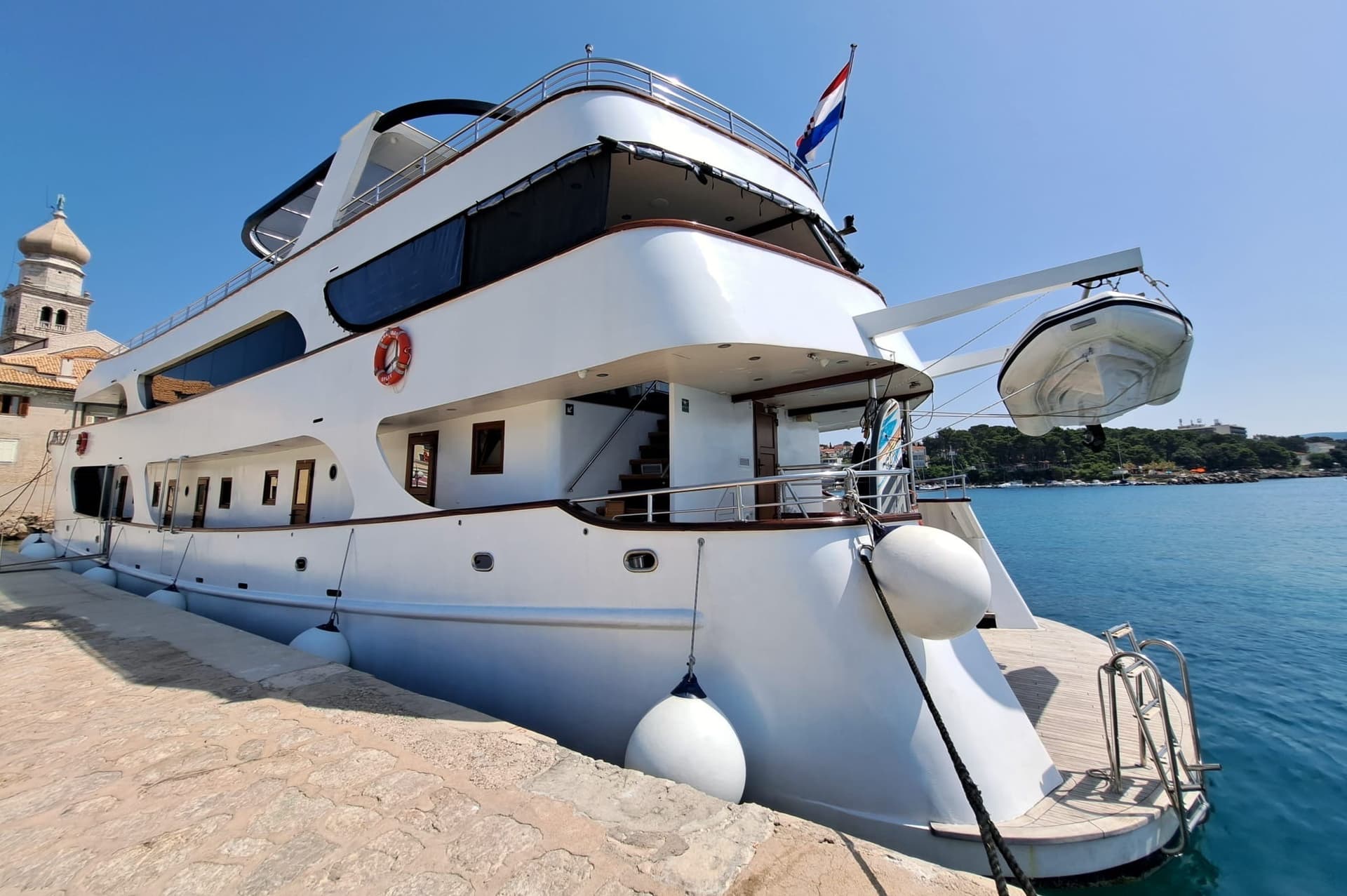 Large white yacht docked beside stone pier with Croatian flag and coastal town in background.