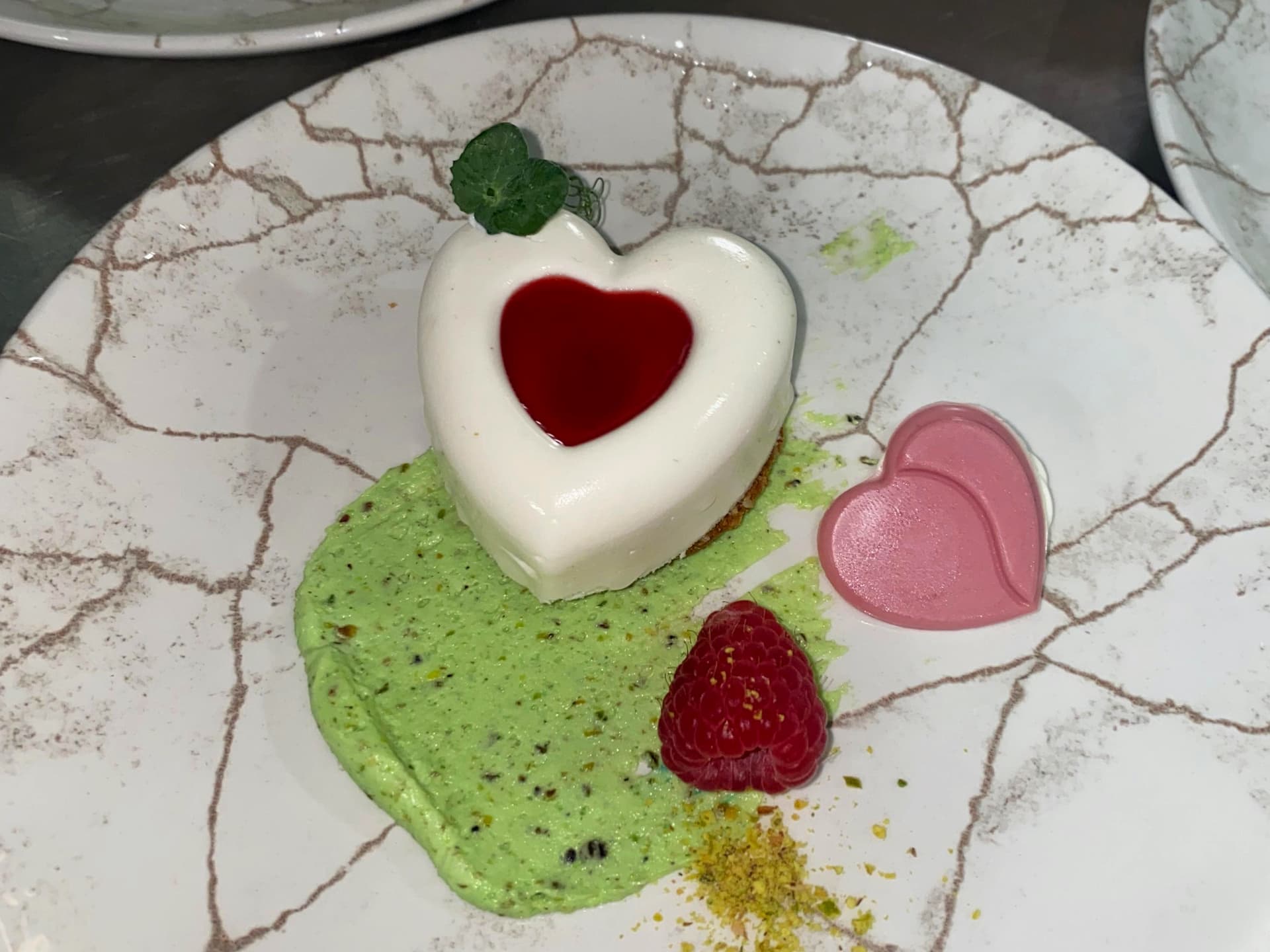 Heart-shaped white dessert with red center, pistachio cream, raspberry, and pink candy on cracked white plate.