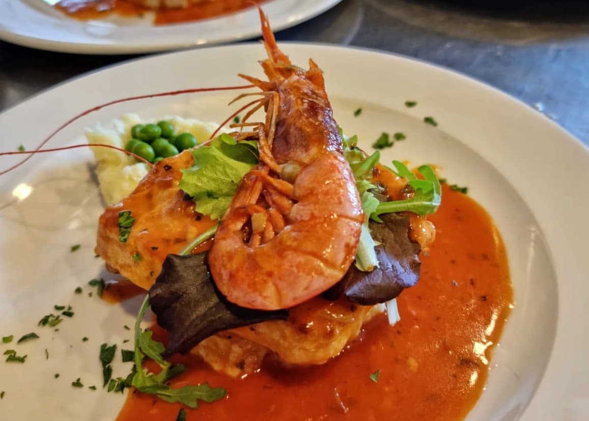 Seafood dish with whole prawn, mashed potatoes, peas, and red sauce on a white plate.