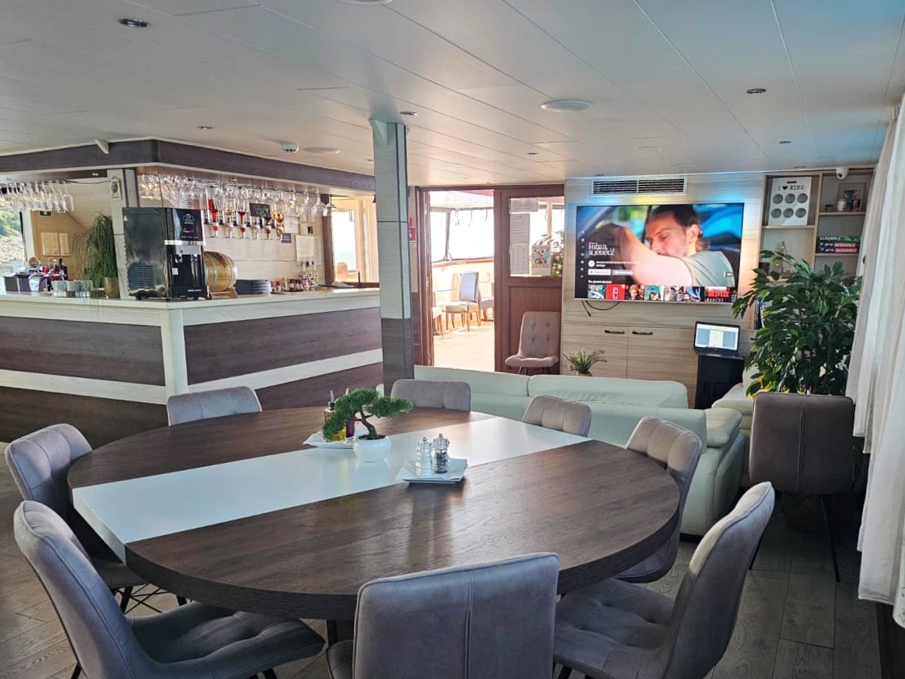 Lounge area with dining table, bar, and TV screen, possibly on a boat or cruise interior.