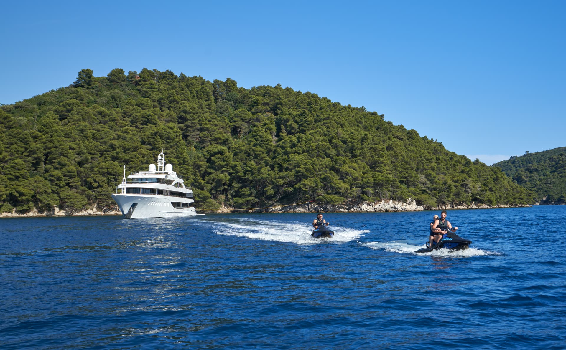 Two people jet skiing on blue water near a large white yacht and forested coastline.