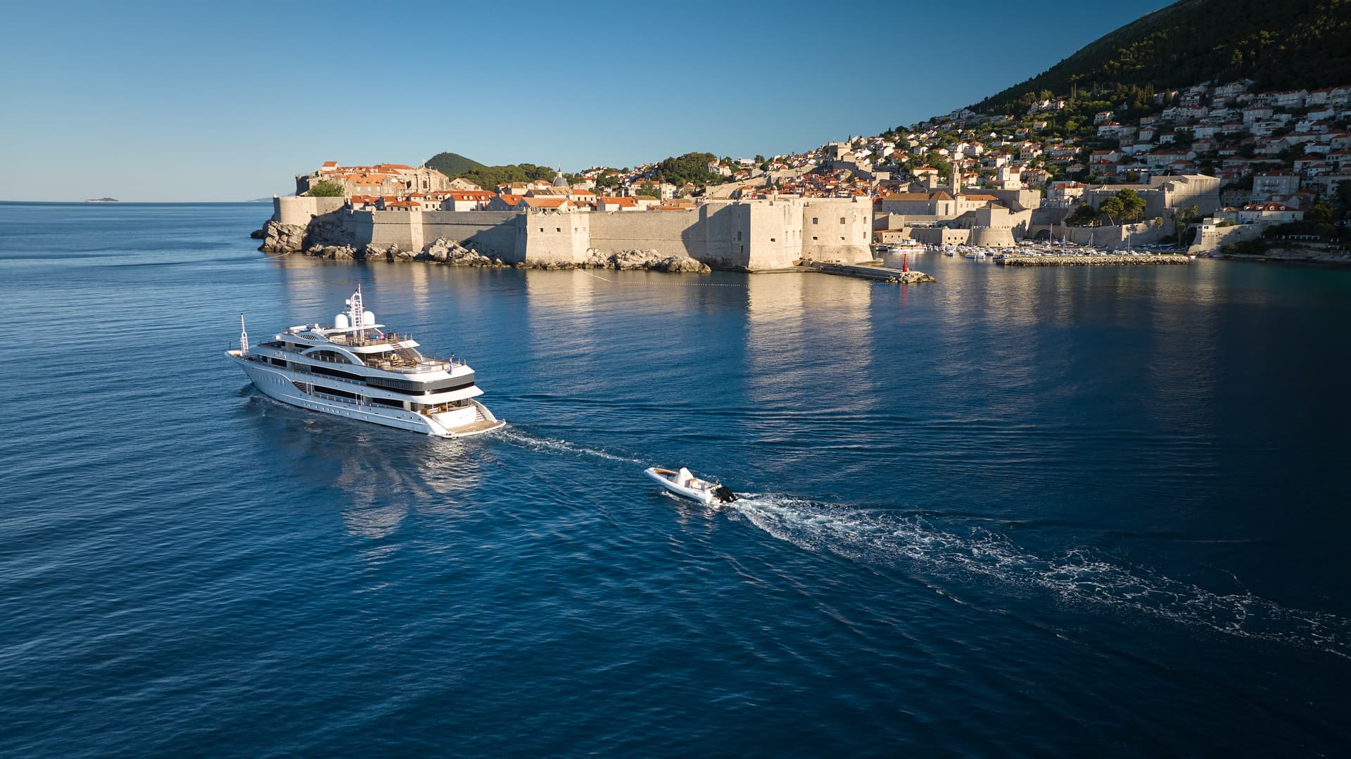 Large white yacht and small tender boat on deep blue sea near walled historic coastal city.
