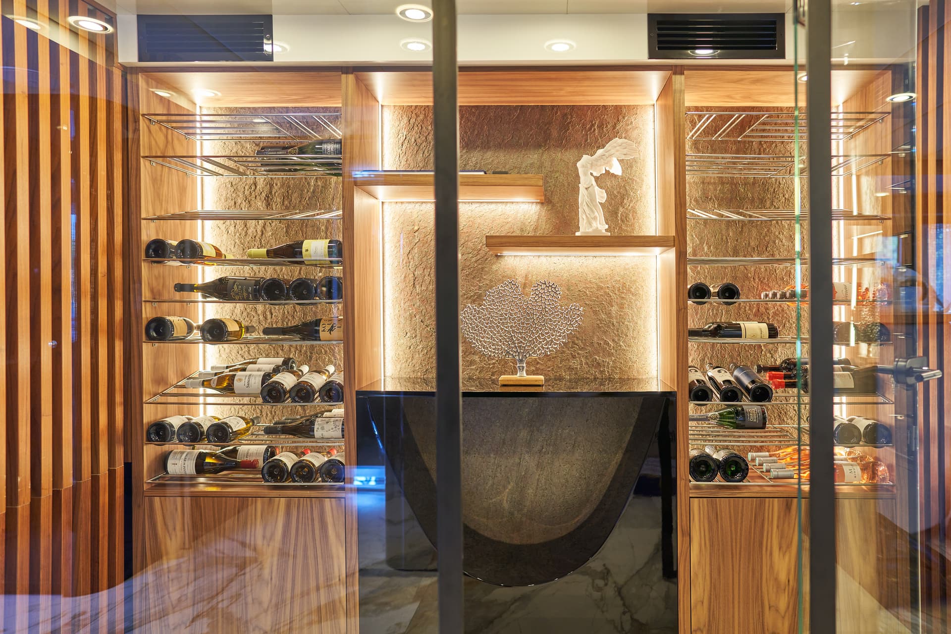Illuminated wooden wine cellar with bottles, marble counter, and decorative statue visible through glass.