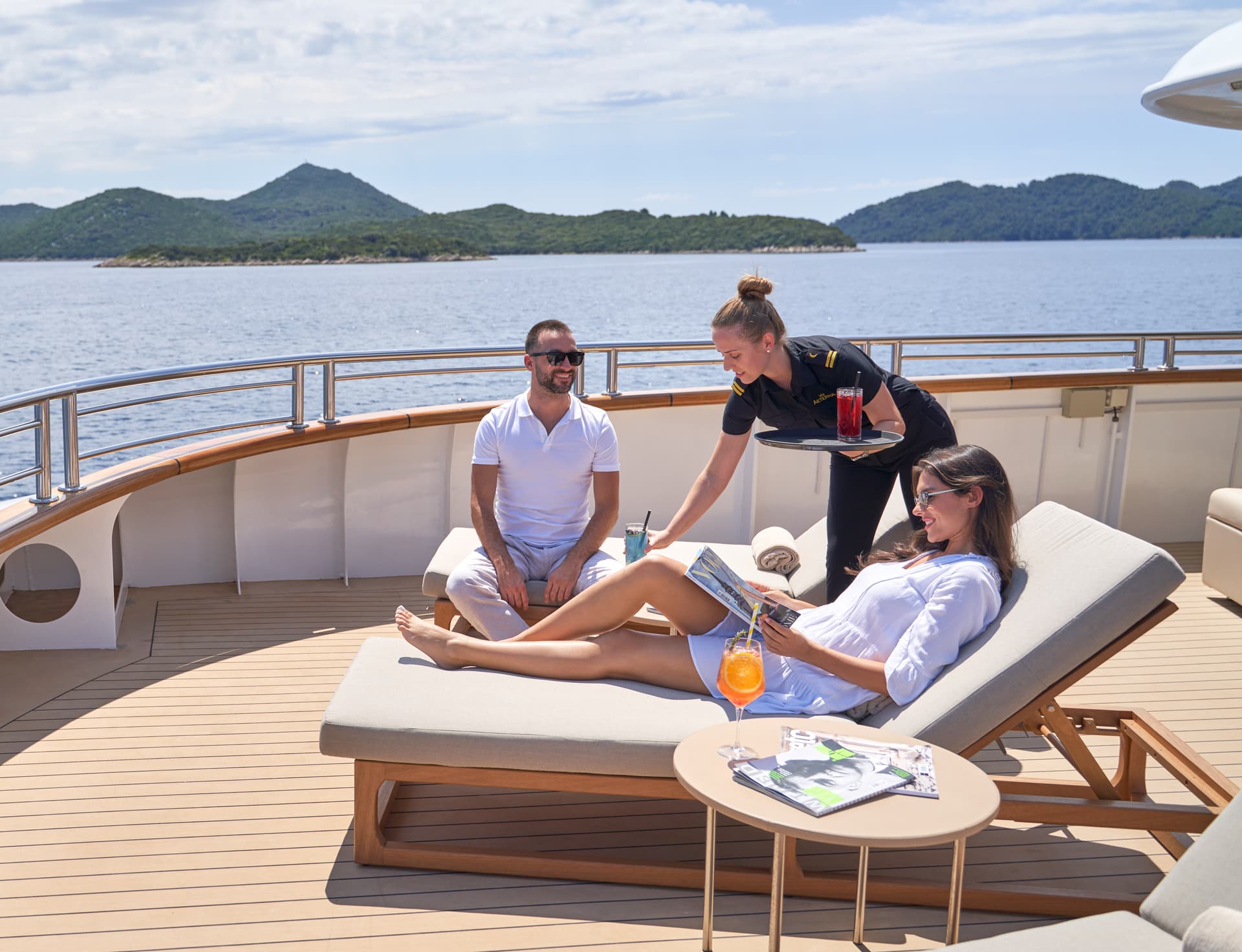 Guests relaxing on yacht deck while crew serves drinks near forested islands on sunny day.