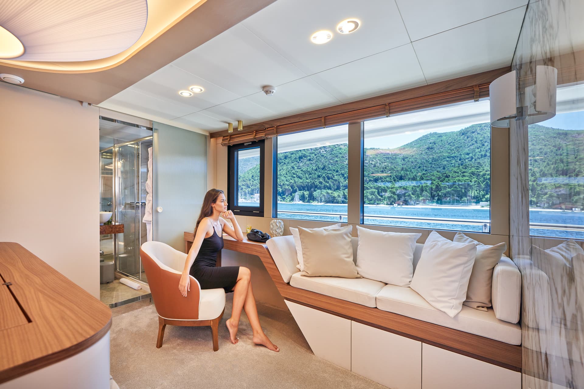 Woman relaxing in modern yacht cabin overlooking lush green coastline and blue water.