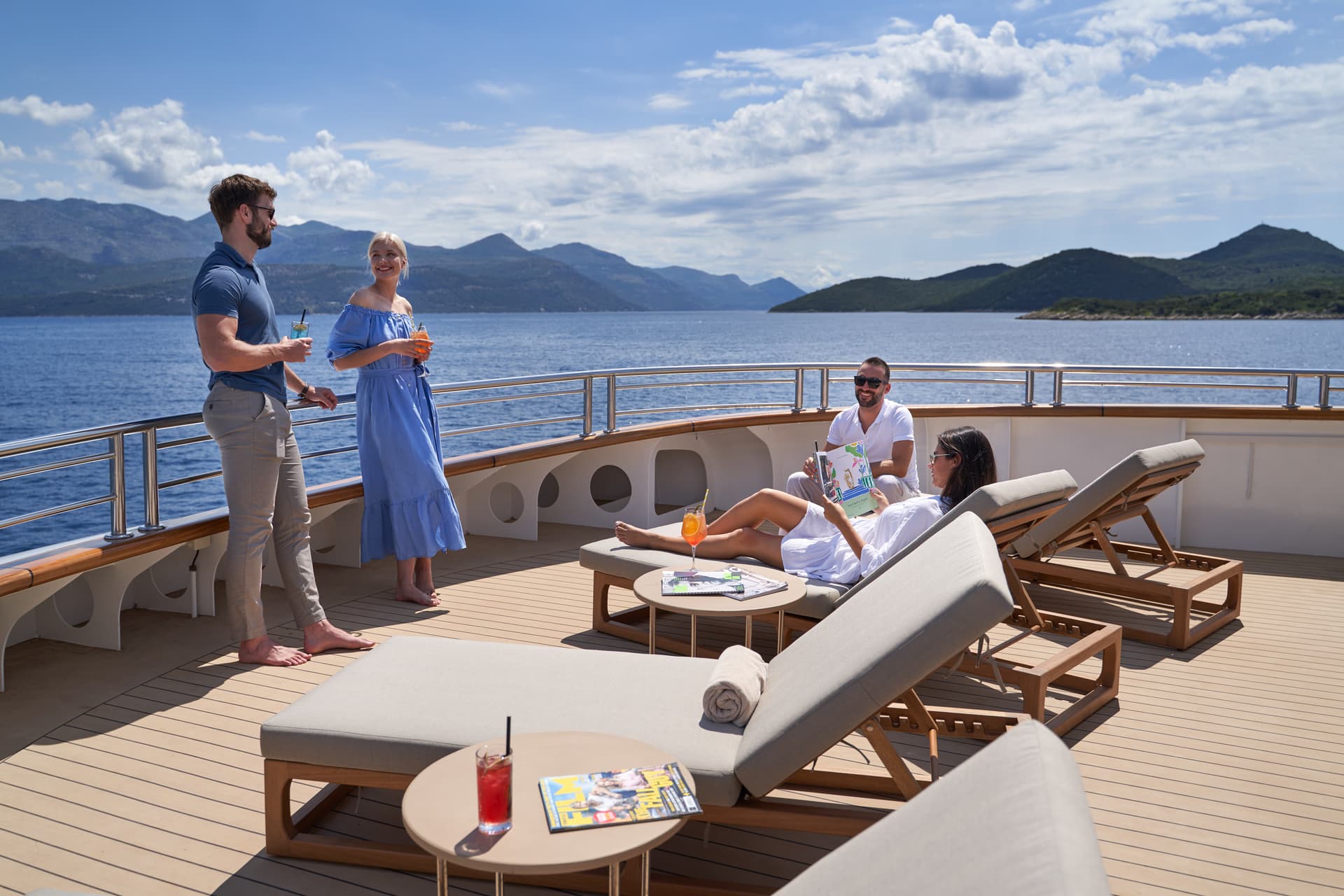 People relaxing on yacht deck with cocktails overlooking mountains and blue sea