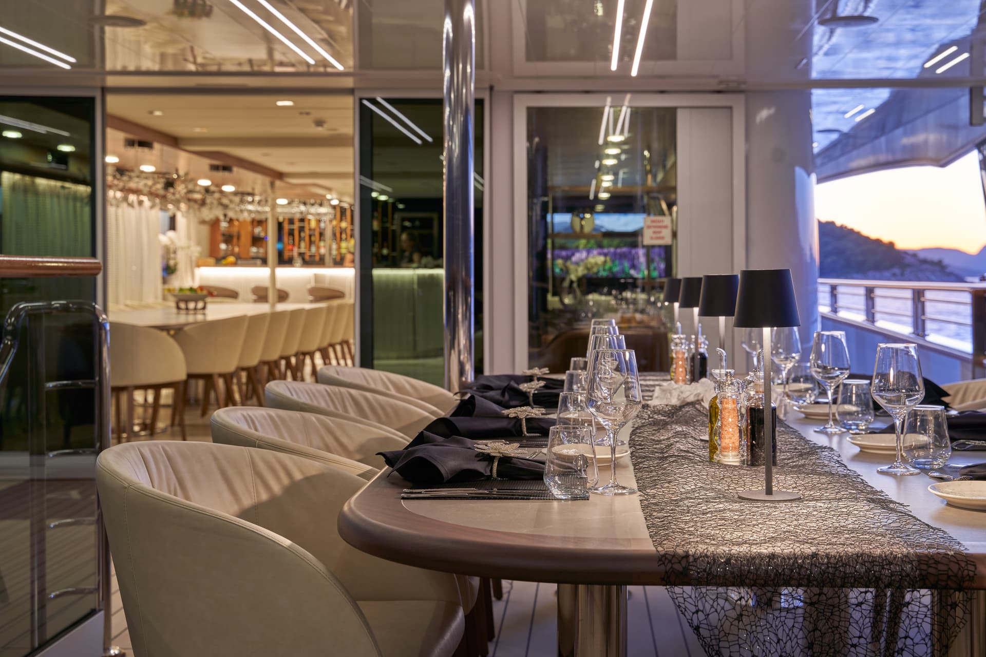Dining table set on a yacht deck overlooking a sunset over coastal mountains.