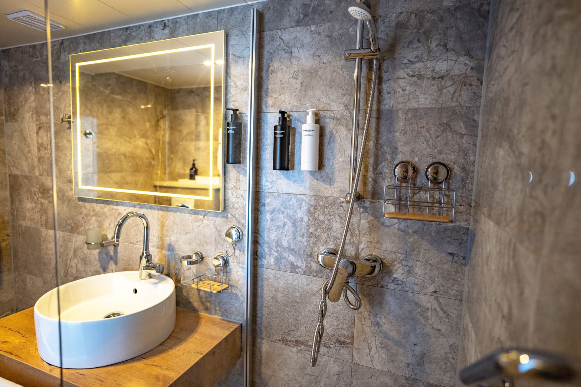 Modern bathroom with vessel sink, illuminated mirror, and chrome shower fixtures against gray marble tile.