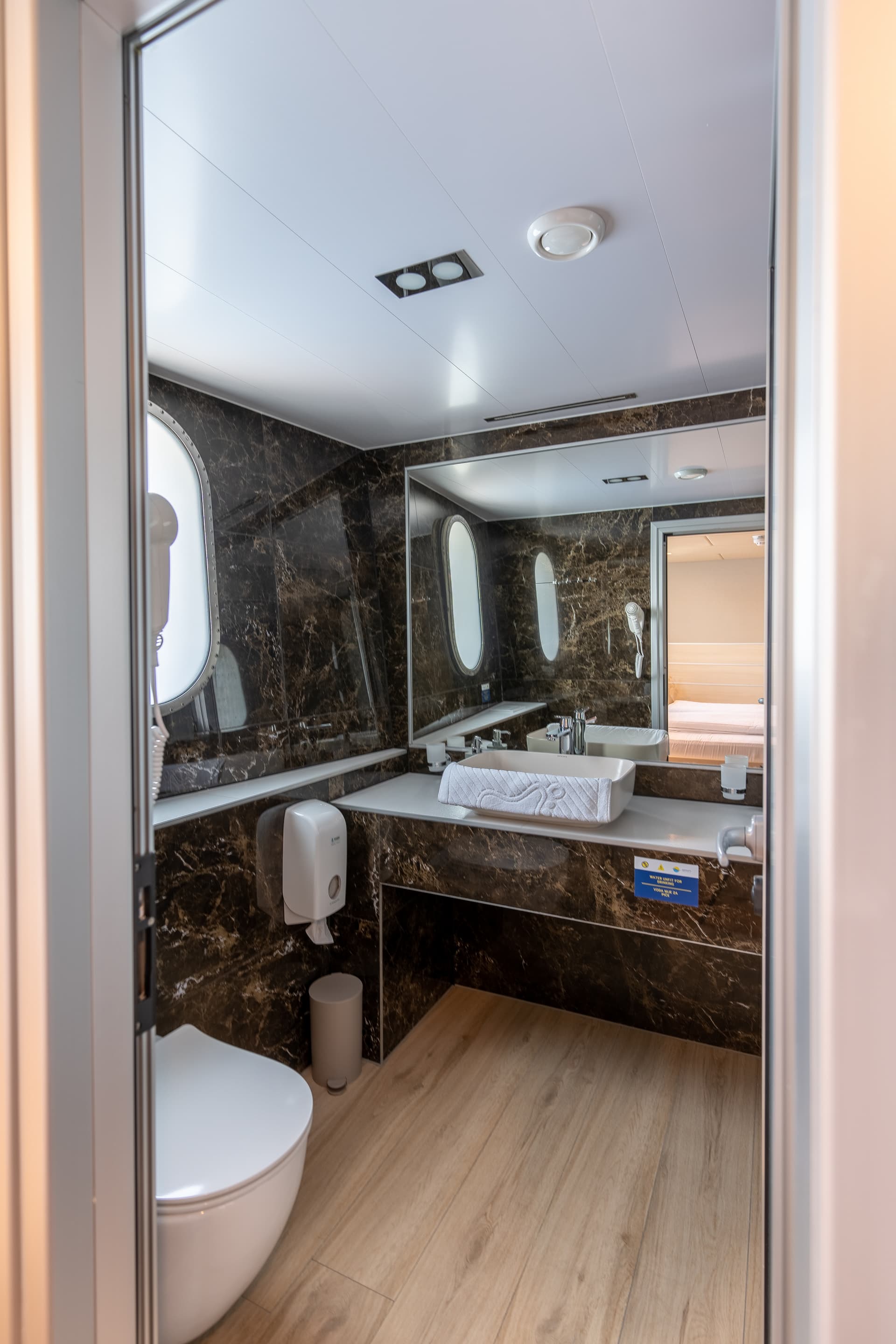 Bathroom with dark marble walls, vessel sink, toilet, and porthole windows suggesting a boat or cruise ship.