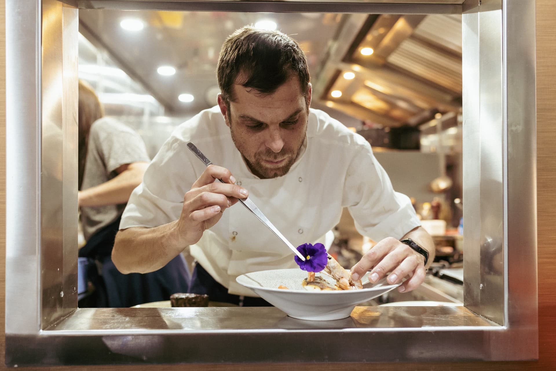 Chef plating seafood dish with tweezers and a purple edible flower in a professional kitchen.