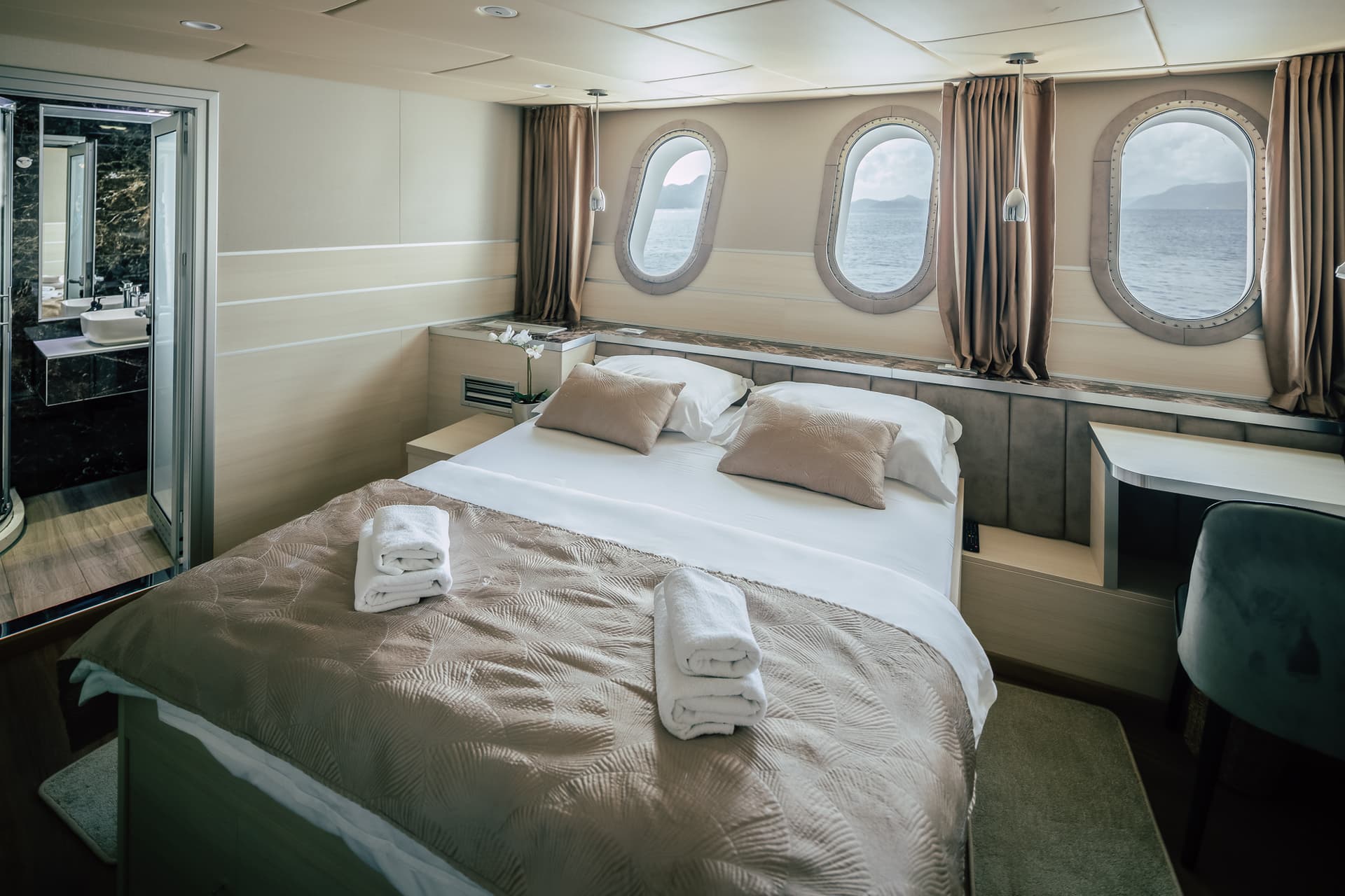 Cabin bedroom with double bed, porthole windows showing sea and mountains, and marble bathroom entrance.