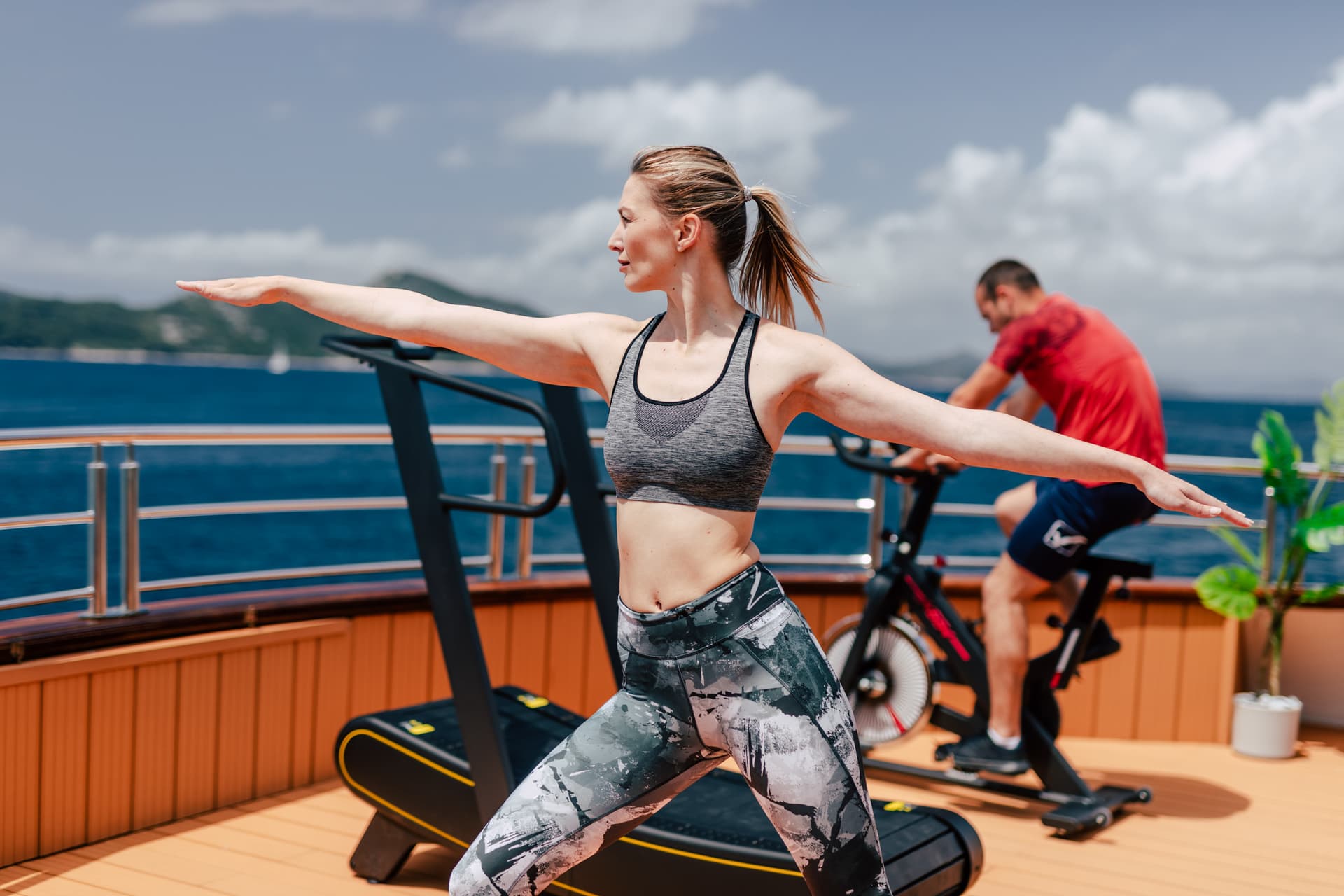 Woman doing yoga pose on deck with ocean view; man cycling nearby.