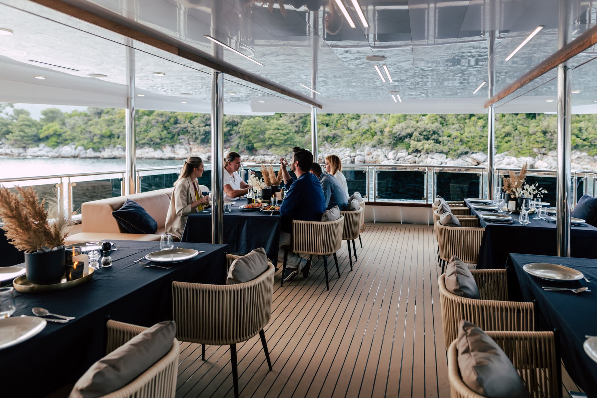Dining on a yacht deck with views of a lush, rocky coastline and calm water.