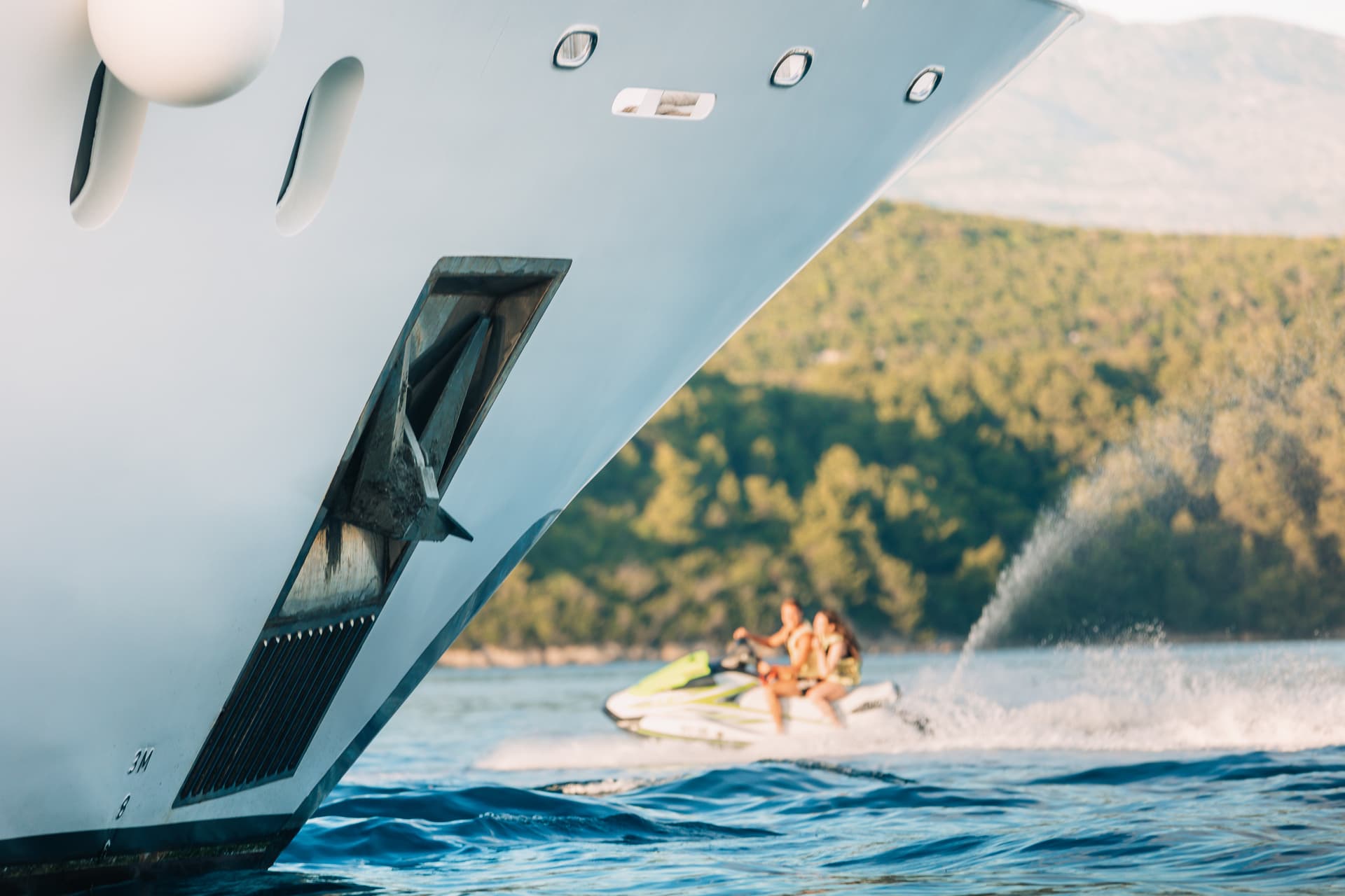 Close-up of yacht bow with jet ski activity on blue water near green coastline.