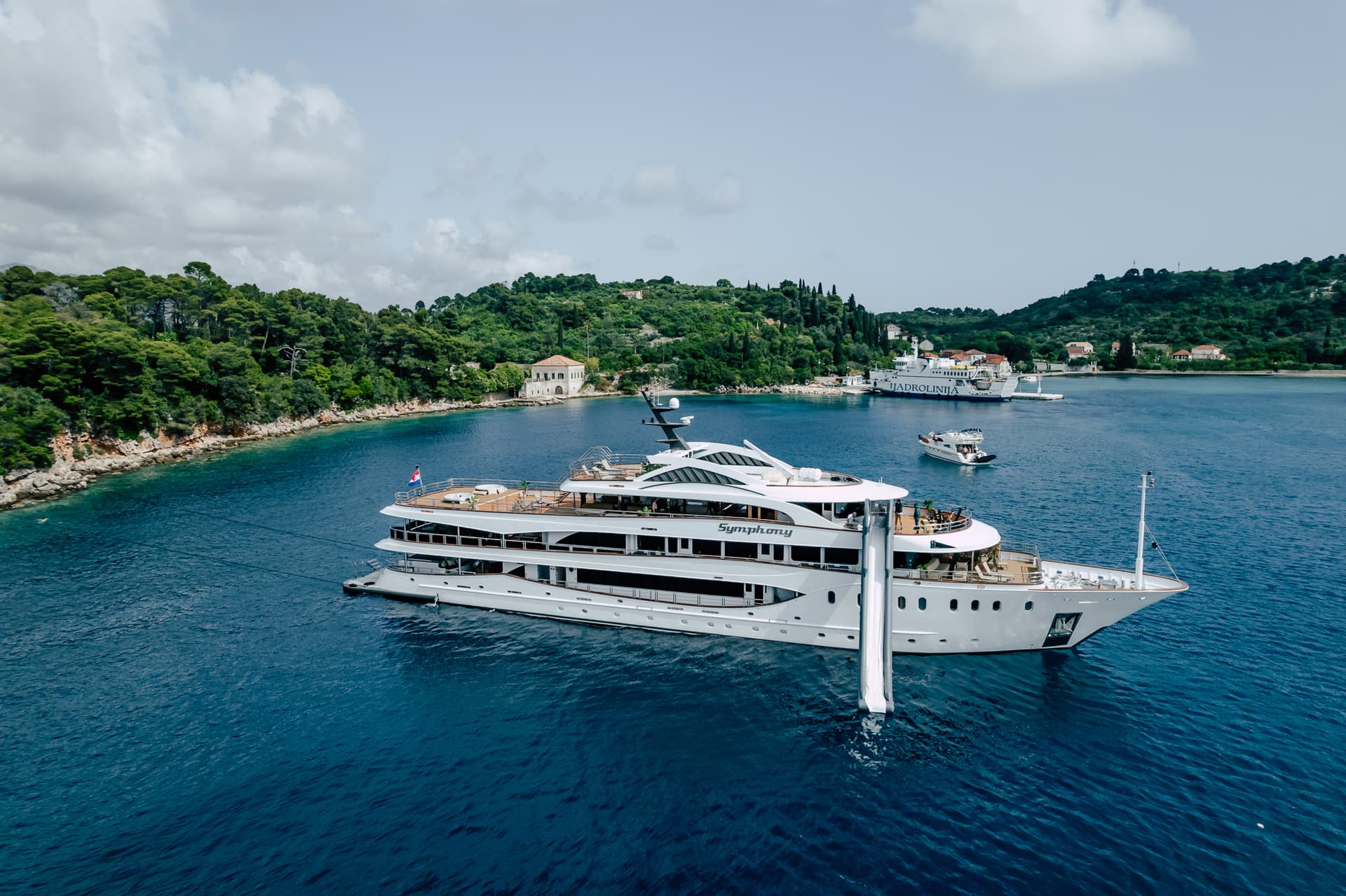 Large white yacht named Symphony with water slide anchored in blue coastal waters near green hills.