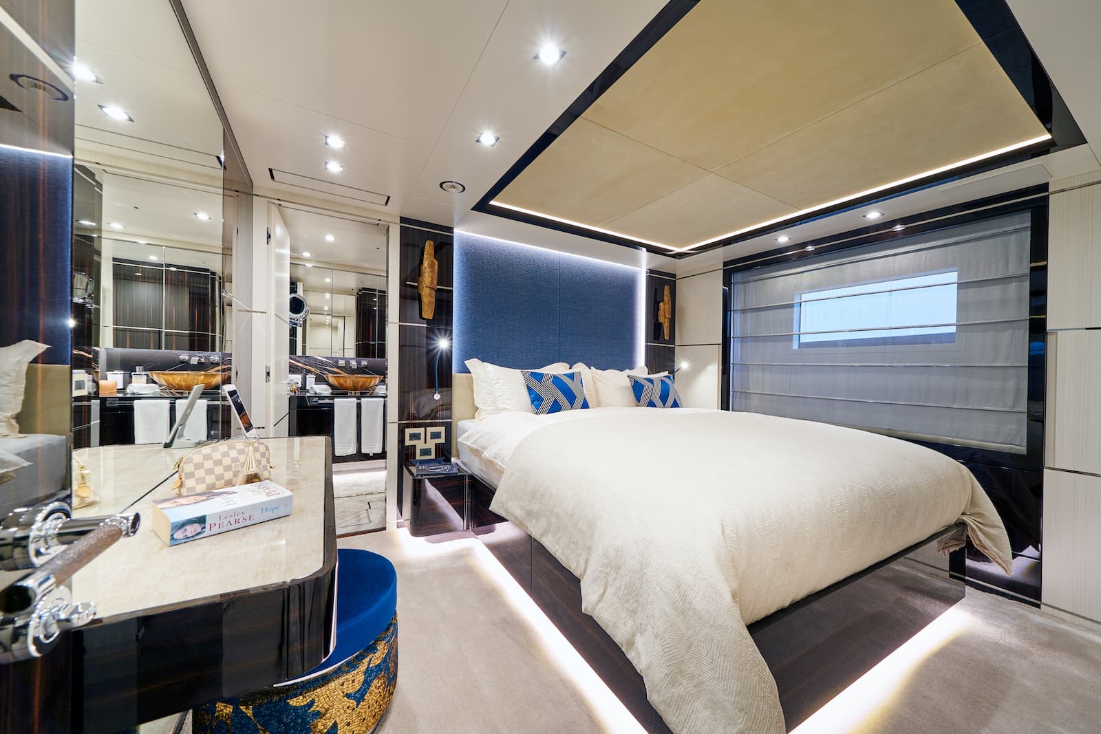 Luxurious yacht stateroom with white bedding, blue accents, and mirrored en-suite bathroom visible.