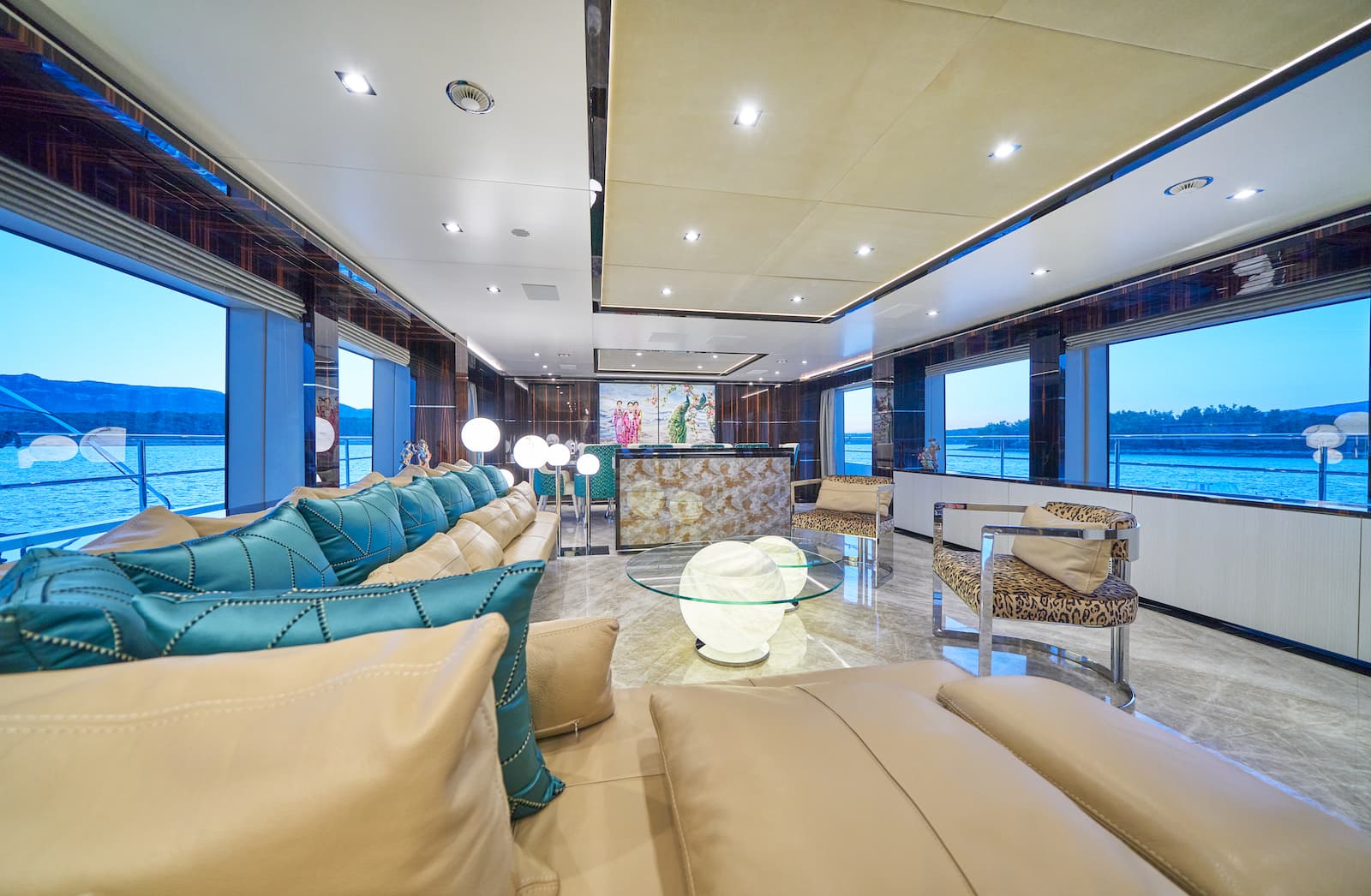 Luxury yacht interior lounge with large windows showing blue water and distant mountains.