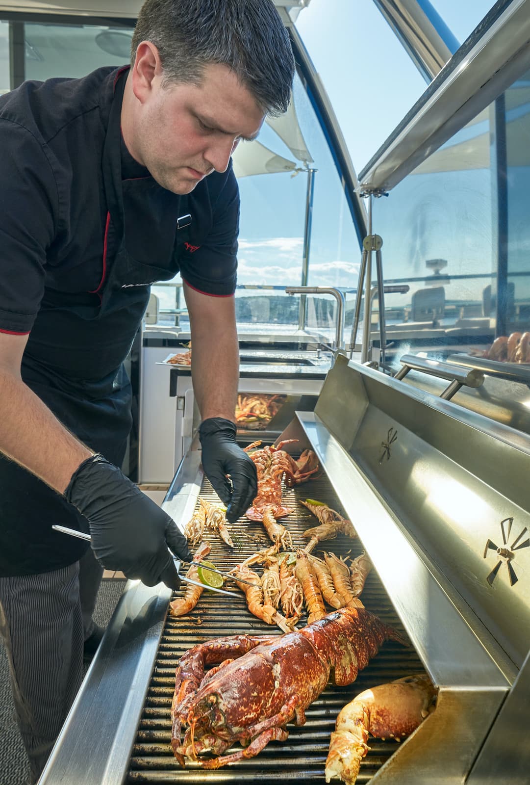 Chef grilling lobster and shrimp with lime slices on an outdoor boat grill.