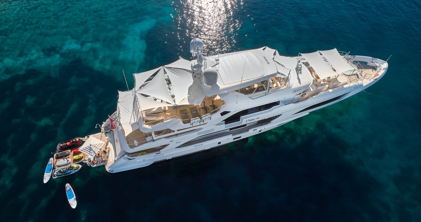 Large white motor yacht anchored in clear turquoise water with sun glare and water toys.