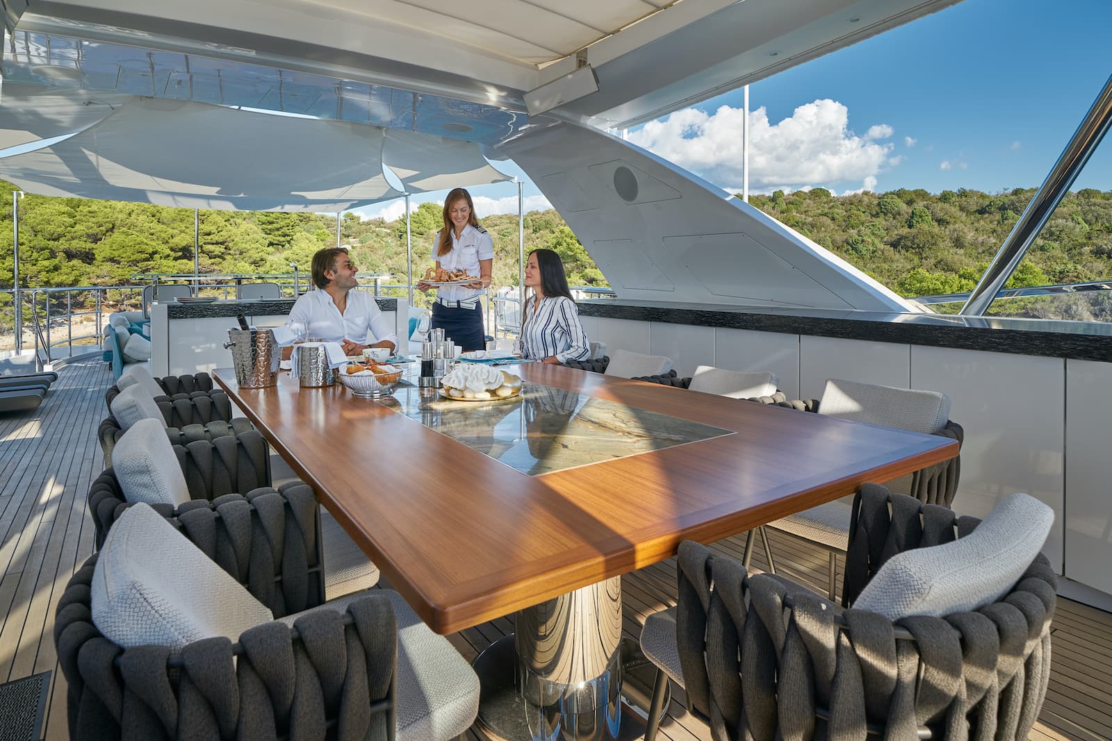 Dining on a yacht deck with crew serving a couple near a lush, green coastline.