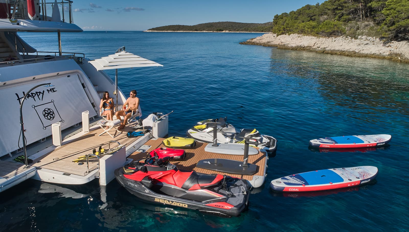 Yacht swim platform with jet skis and paddleboards anchored near a wooded coastline.