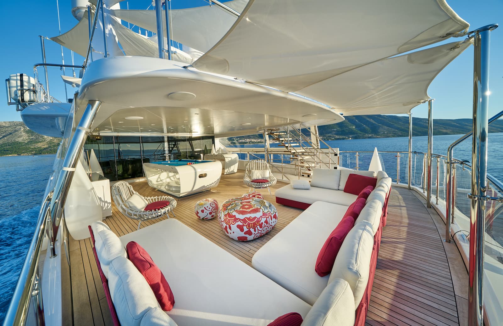 Luxury yacht deck with white lounge seating, pool table, and mountainous coastline view.