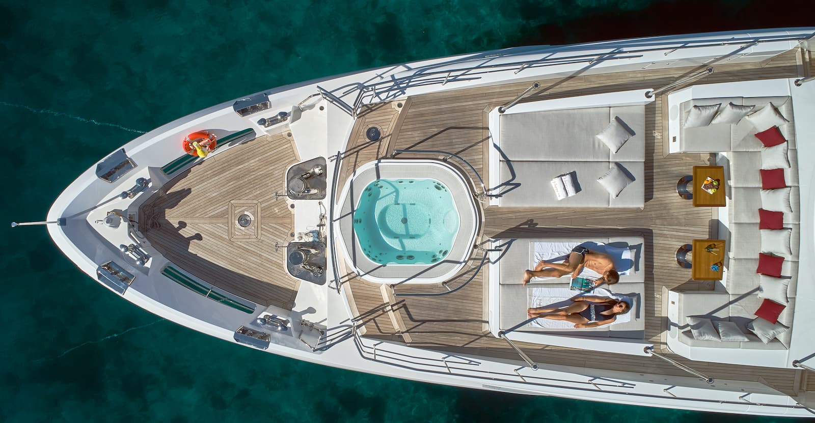 Aerial view of yacht deck with jacuzzi, sun loungers, and people relaxing on turquoise water.