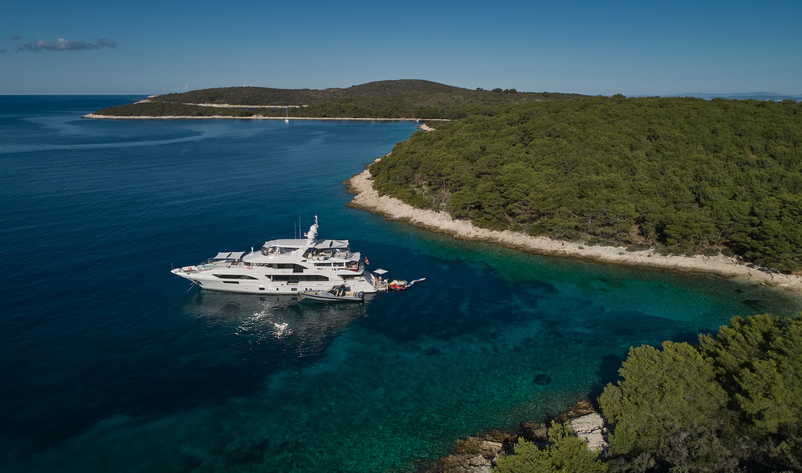 Luxury yacht anchored in clear turquoise cove near forested coastline under blue sky.