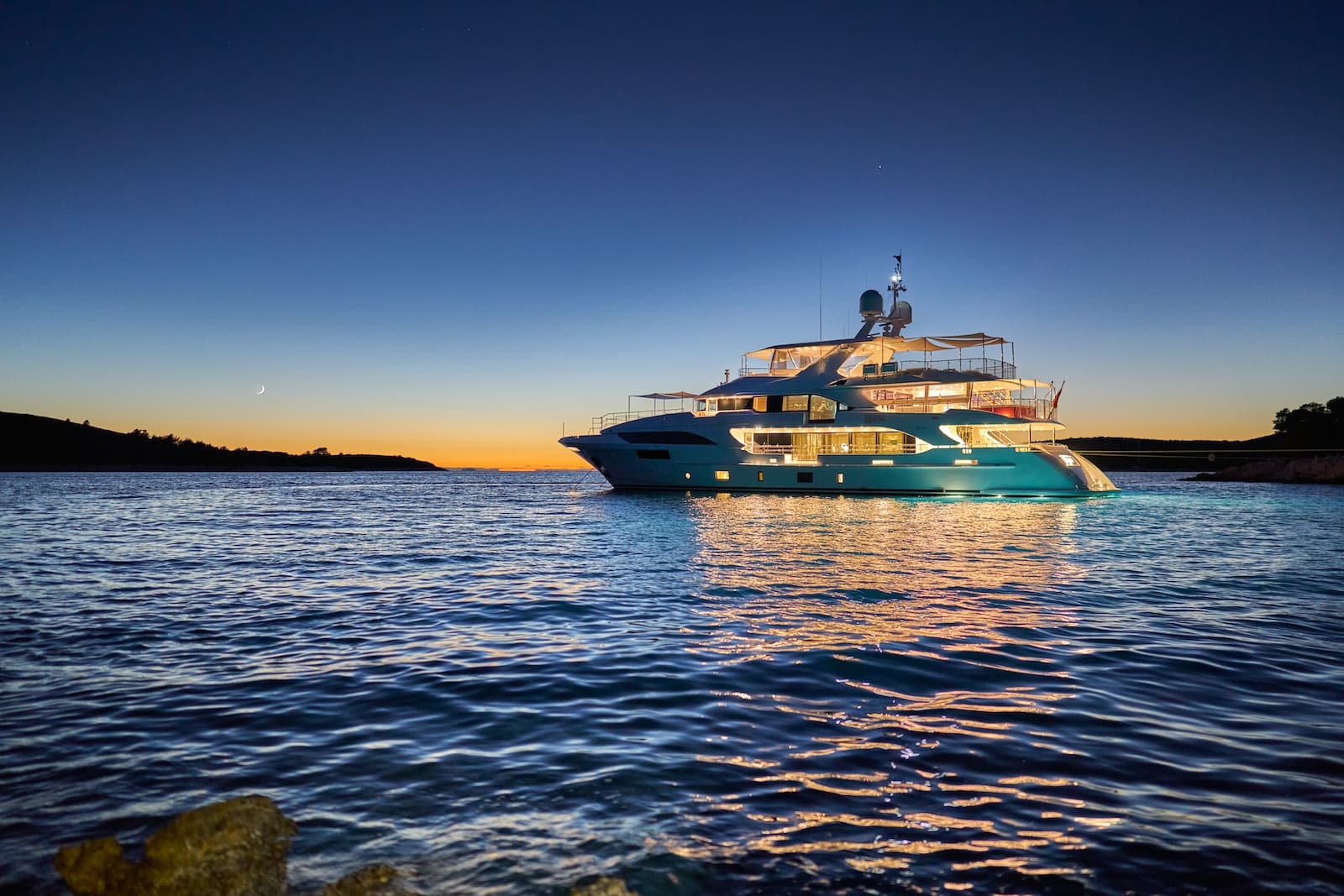 Luxury yacht illuminated at anchor on dark water near a coastline at twilight with a crescent moon.