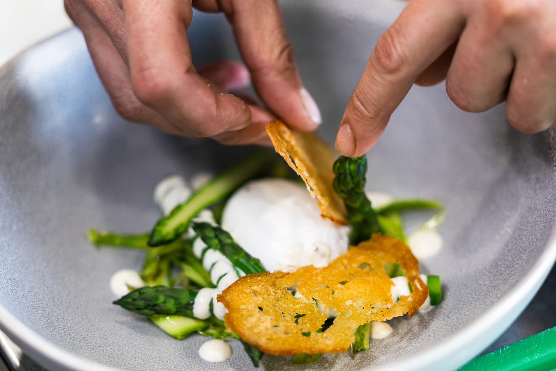 Chef garnishing poached egg with asparagus, crisp bread, and white sauce in a gray bowl.